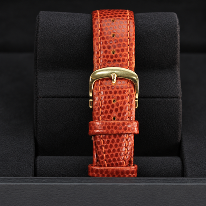 A textured orange-brown leather watch strap with a gold buckle, displayed vertically on a black watch cushion.