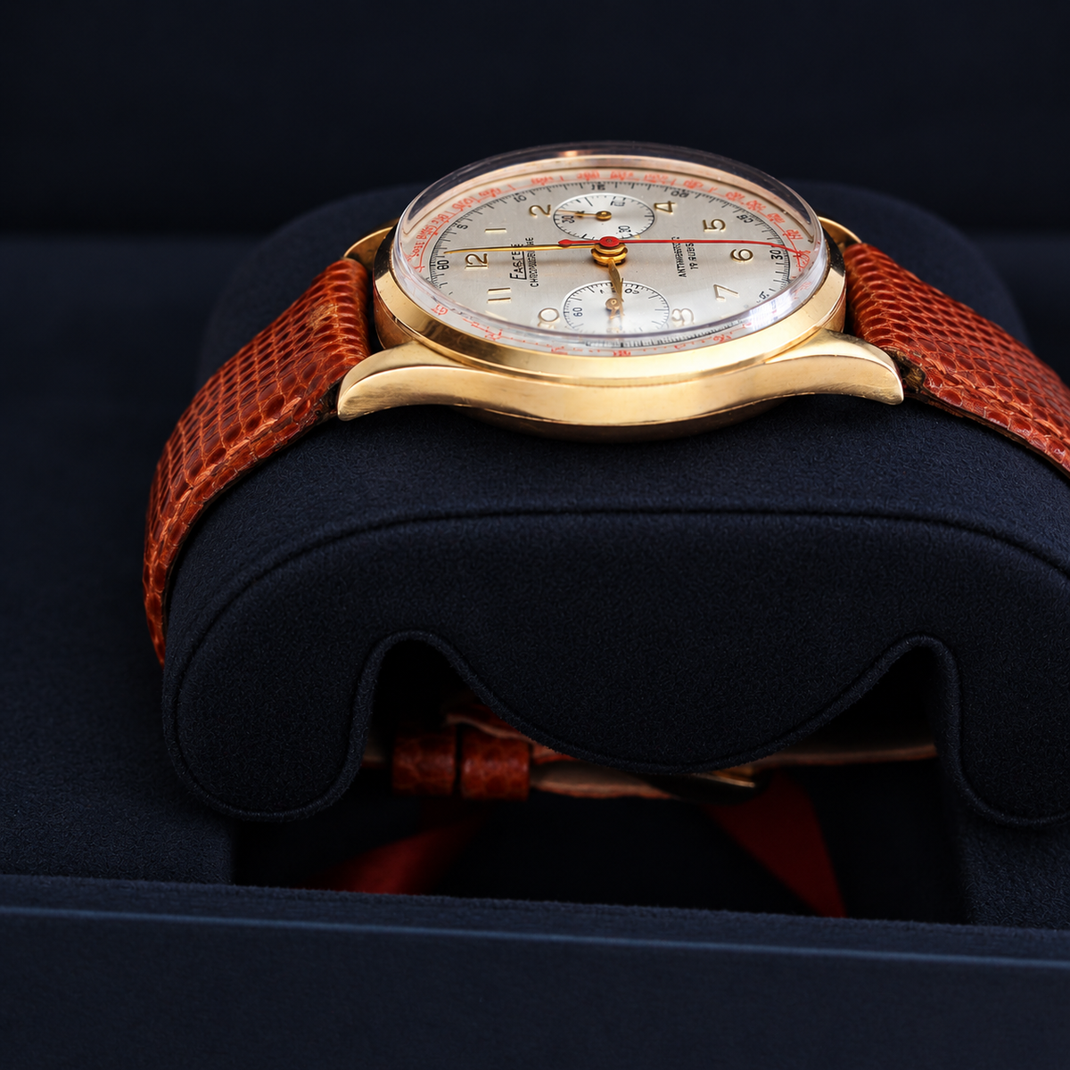 A vintage-style wristwatch with a gold-tone case, white dial, and textured reddish-brown strap, displayed on a dark cushion inside a presentation box.