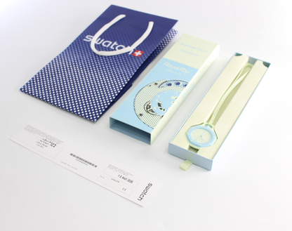 Swatch watch packaging and accessories on a white background, including a blue patterned Swatch shopping bag, a light blue and white Swatch box, a pale green watch strap, and a watch in a pale green presentation box.