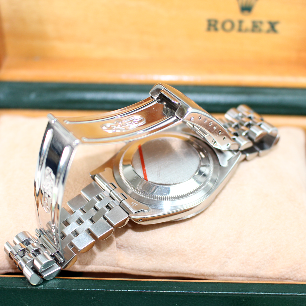 Close-up of a silver watch with a detailed band on a beige cushion, with a Rolex box in the background.