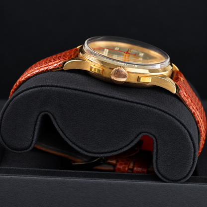 A close-up side view of a vintage-style wristwatch with a gold-toned case, clear crystal, and textured orange-brown leather strap, resting on a black display cushion.