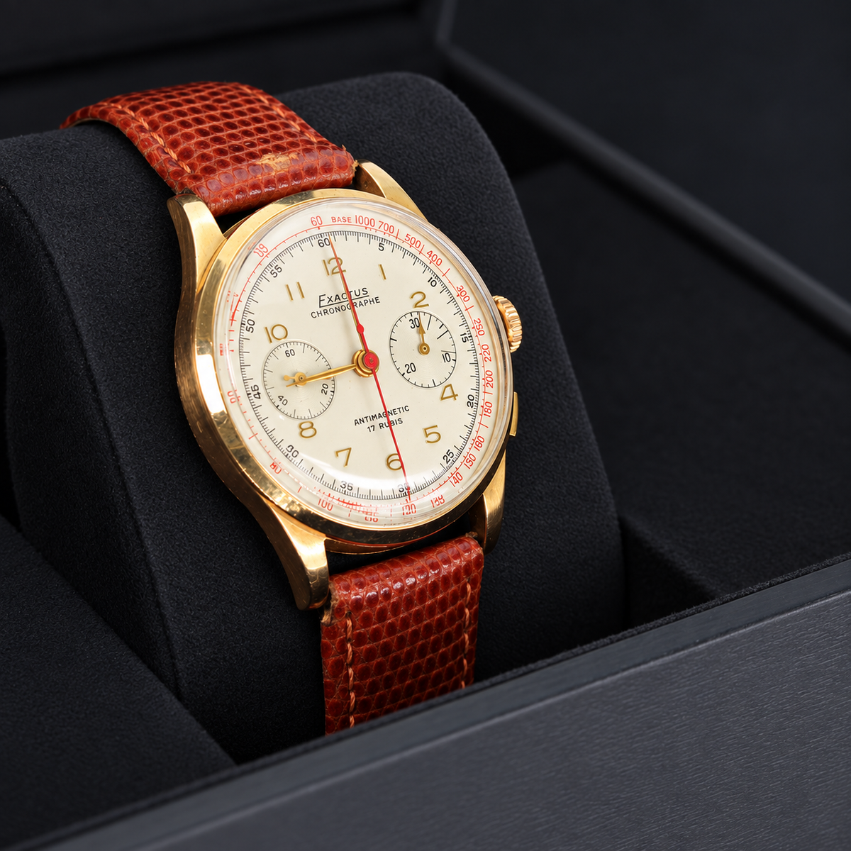 A close-up of a vintage-style wristwatch with a gold-tone case, white dial, and reddish-brown textured strap, displayed inside a black watch box.