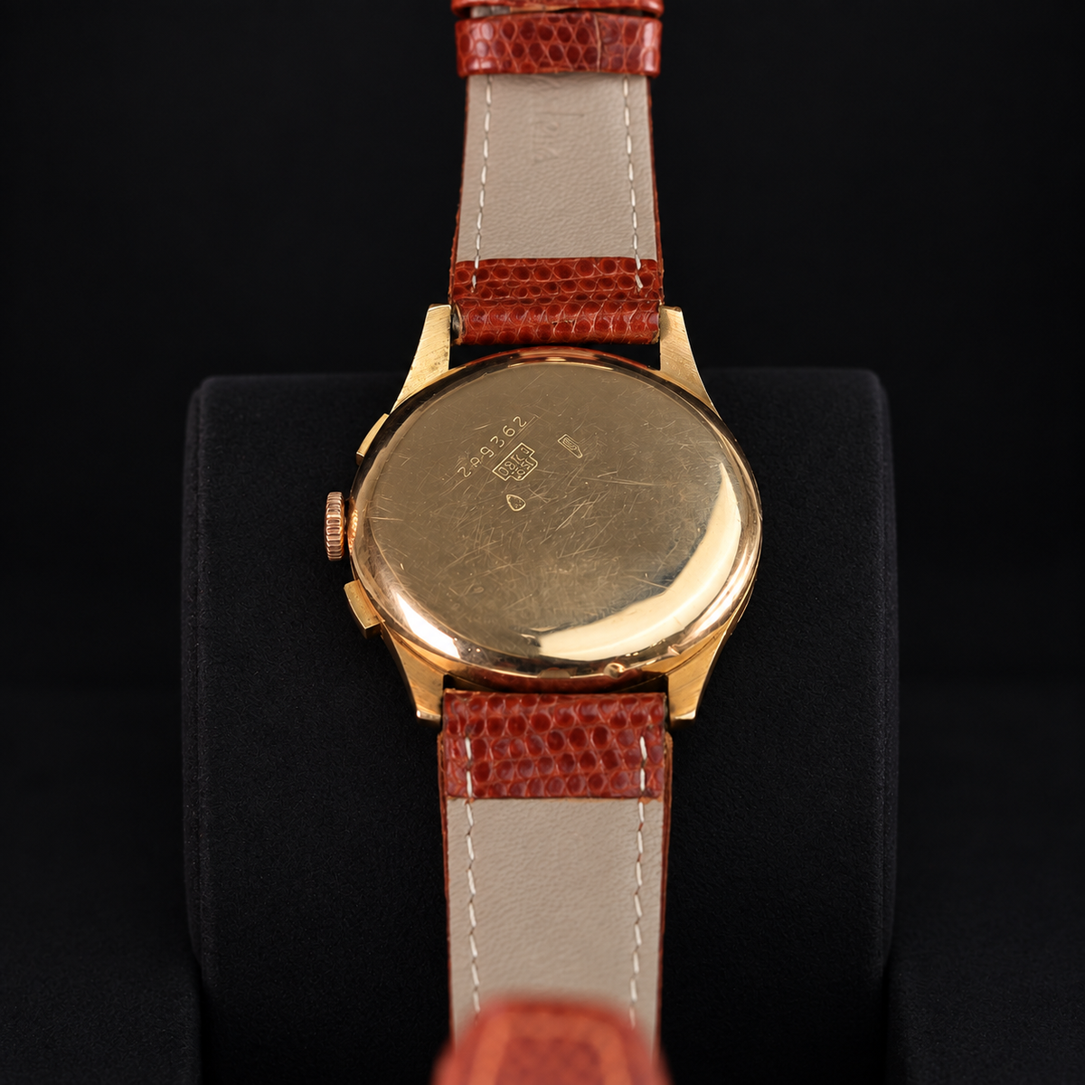 A vintage wristwatch shown from the back, featuring a gold-toned metal case and a reddish-brown textured leather strap with a light beige center stripe, displayed on a black stand against a black background.