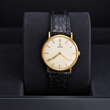 An Omega wristwatch with a gold case, cream dial, and textured black leather strap, displayed on a black cushion inside a watch box.