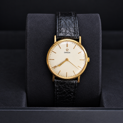 An Omega wristwatch with a gold case, cream dial, and textured black leather strap, displayed on a black cushion inside a watch box.