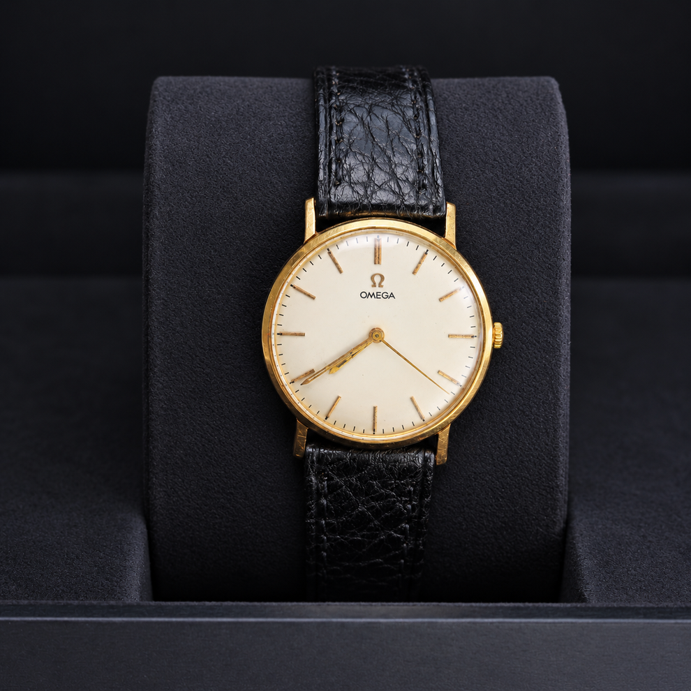 An Omega wristwatch with a gold case, cream dial, and textured black leather strap, displayed on a black cushion inside a watch box.