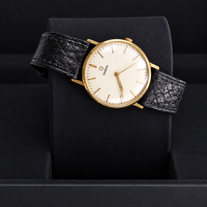 Omega wristwatch with a gold-tone case, cream dial, and textured black leather strap, displayed on a black cushion.