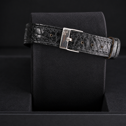 A black textured leather watch strap with a silver-toned metal buckle, displayed on a black watch cushion.