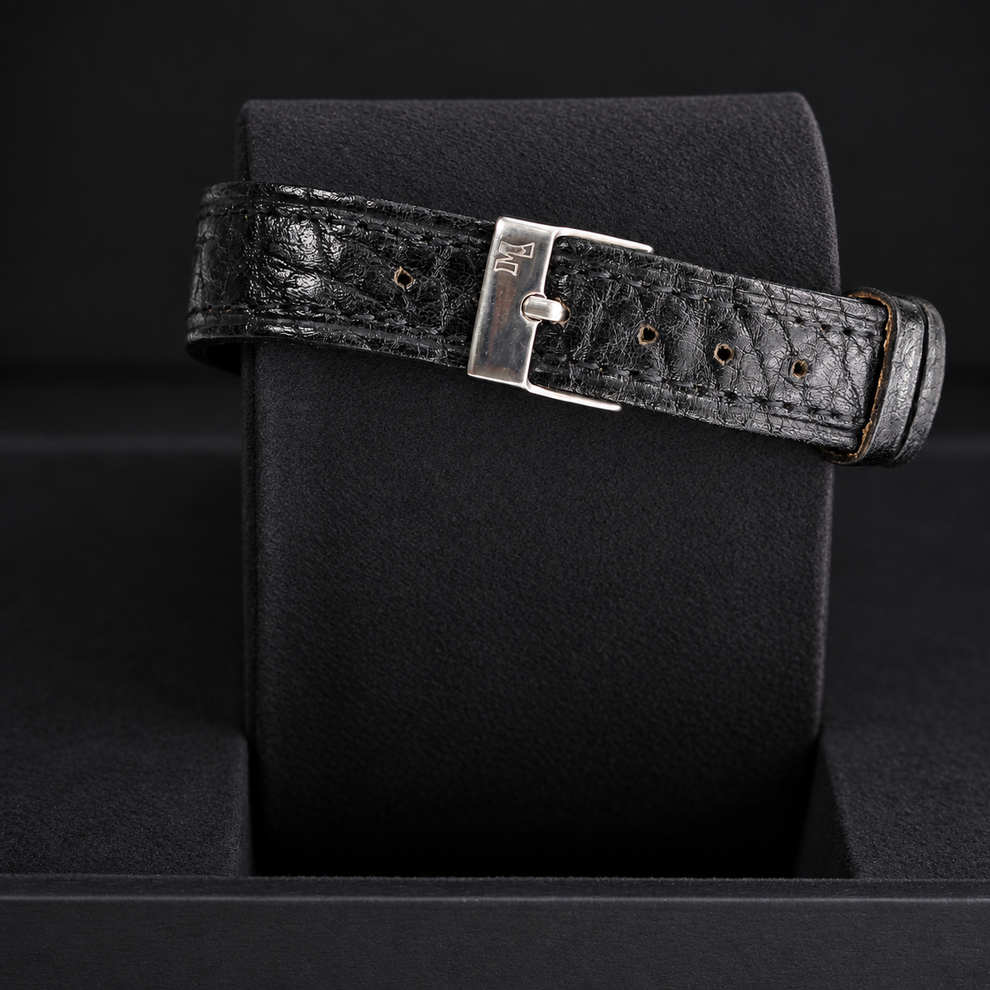 A black textured leather watch strap with a silver-toned metal buckle, displayed on a black watch cushion.