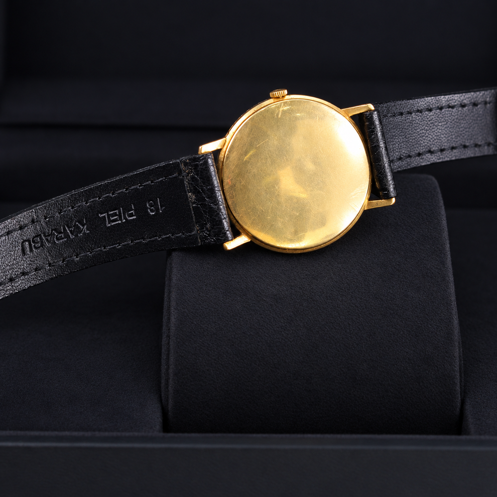 A vintage-style wristwatch with a round gold-toned case and a black leather strap, photographed resting on a black display cushion inside a black presentation box.