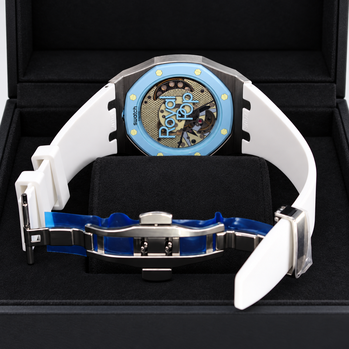 A luxury watch with a white rubber strap, a light blue bezel, and a visible skeleton dial with gold details, presented inside a black watch box.