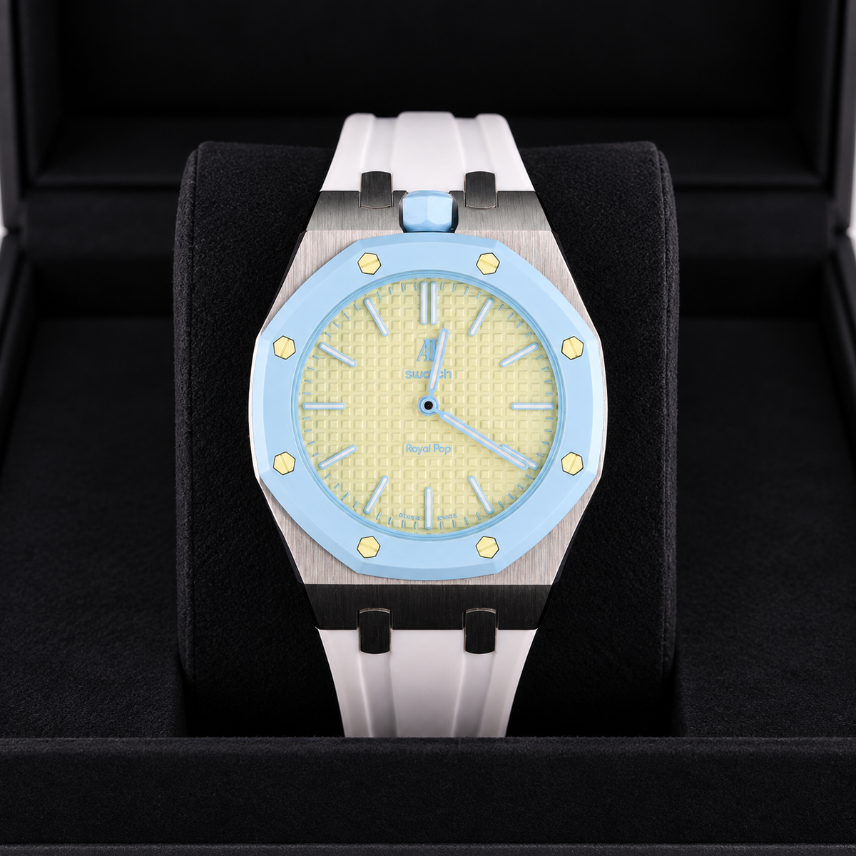 A luxury watch with a light blue bezel, textured pale yellow dial, and a white rubber strap, presented inside a black watch box.