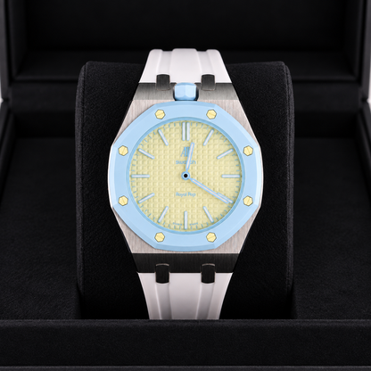 A luxury watch with a light blue bezel, textured pale yellow dial, and a white rubber strap, presented inside a black watch box.