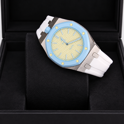 A luxury watch with a light blue bezel, textured pale yellow dial, and a white rubber strap, displayed inside a black presentation box.