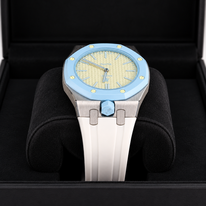A luxury watch with a light blue bezel, a textured pale yellow dial, and a white ceramic strap, presented inside a black watch box.
