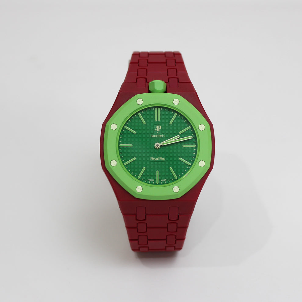 A luxury watch with a deep red metal link bracelet, a green dial, and a green bezel accented with silver-tone screws, shown on a light gray background.
