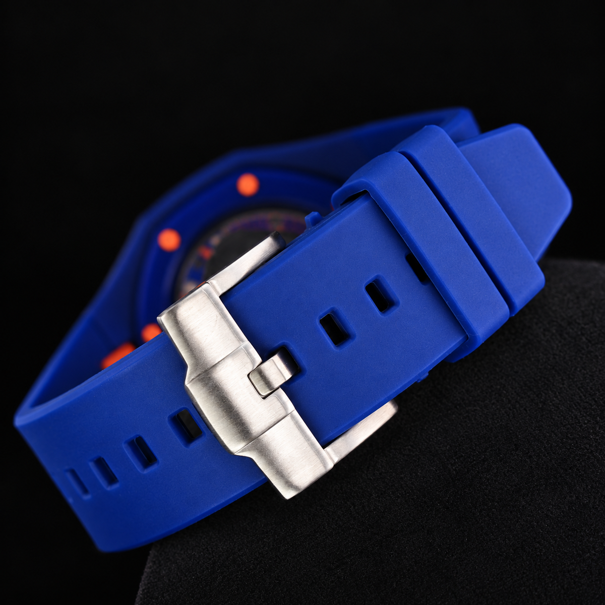 A close-up of a watch with a bright blue silicone strap and a silver metal buckle, set against a dark background.