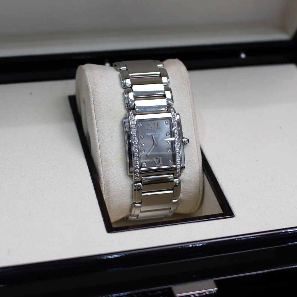 A silver-tone metal link watch with a rectangular face and crystal-accented bezel, displayed on a cream cushion inside a watch box.