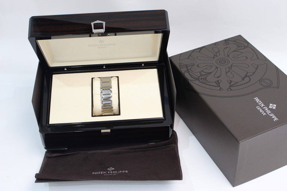 A Patek Philippe watch presented in an open dark wood box with a cream interior, alongside a branded dark brown presentation box and a matching cloth, on a white background.