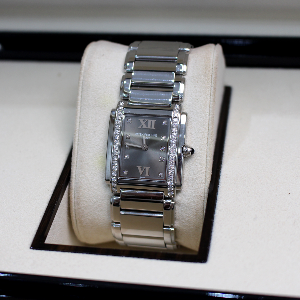 A silver-tone stainless steel Tank watch with a dark gray dial and crystal-embellished bezel, displayed on a cream-colored cushion inside a watch box.