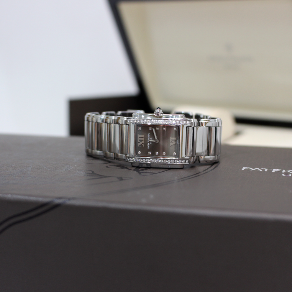 A silver-toned stainless steel watch with a rectangular bezel and integrated link bracelet, resting on a dark gray watch box with its open presentation box blurred in the background.