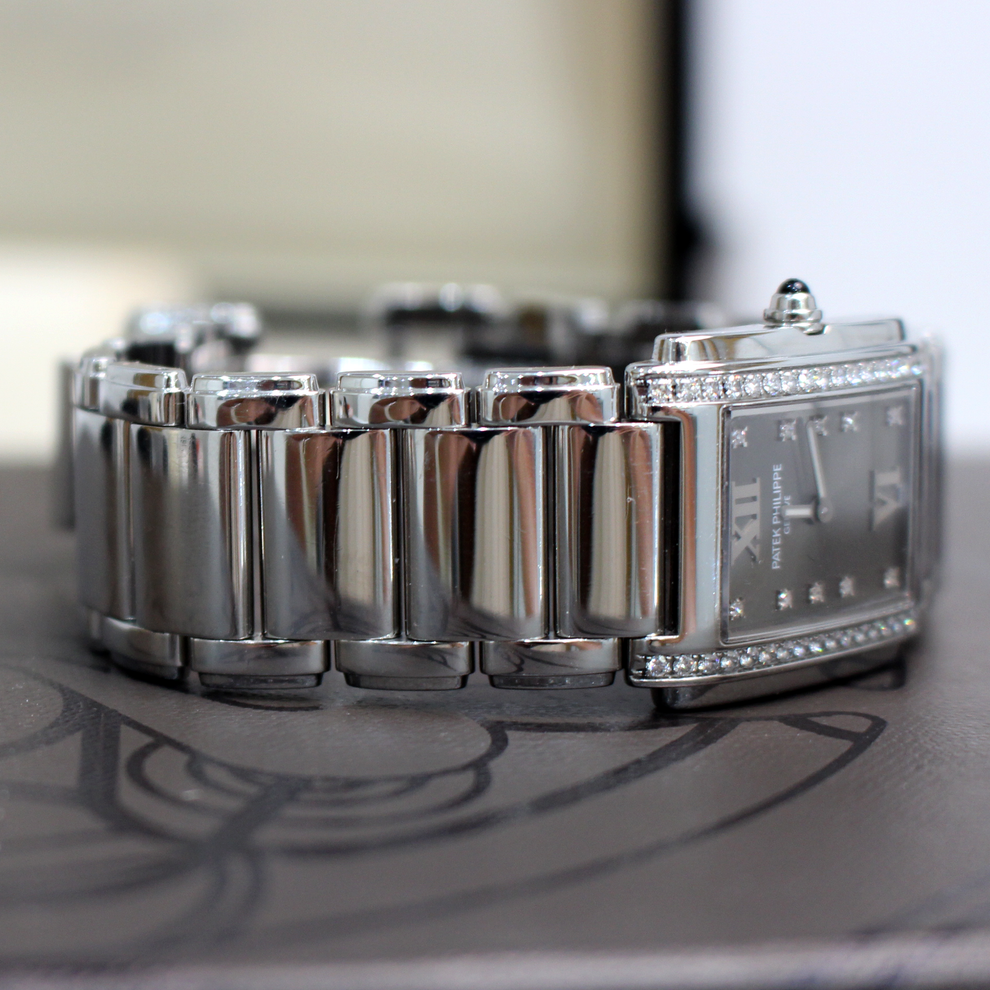 A silver-toned stainless steel tank watch with a polished link bracelet, crystal-accented bezel, and dark dial, resting on a dark surface.