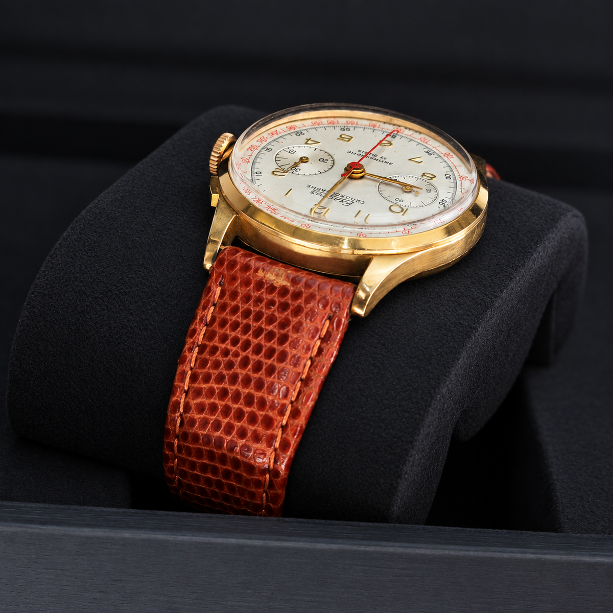 A vintage-style wristwatch with a gold-tone case, white dial, and textured reddish-brown leather strap, displayed on a black cushion inside a watch box.