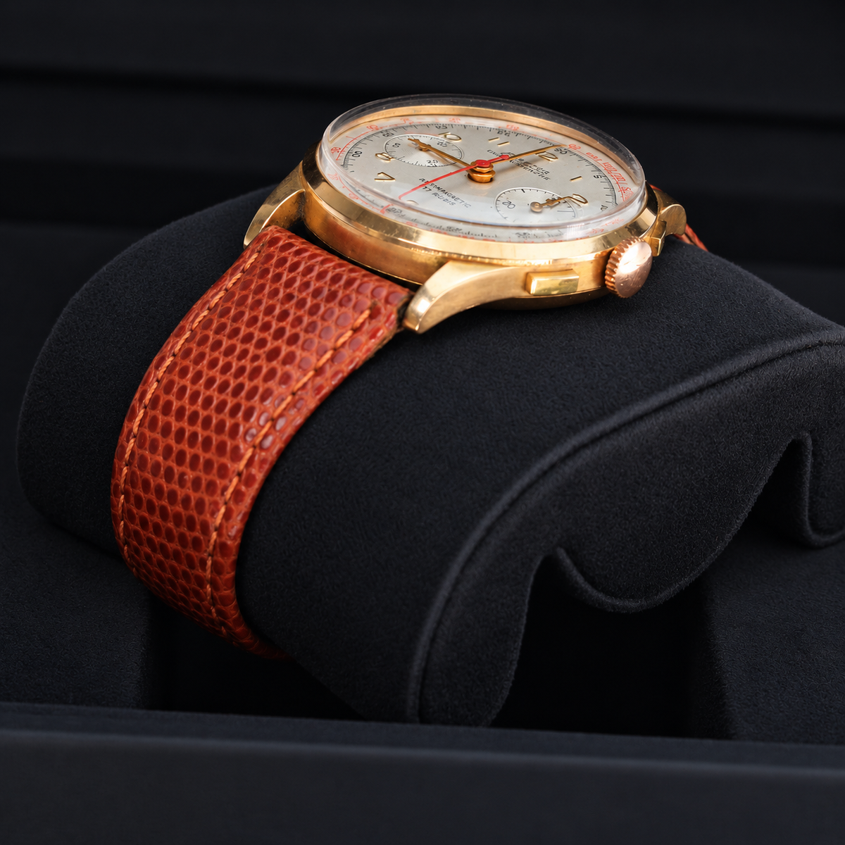A wristwatch with a gold-tone case, cream dial, and textured brown leather strap, displayed on a black cushion.